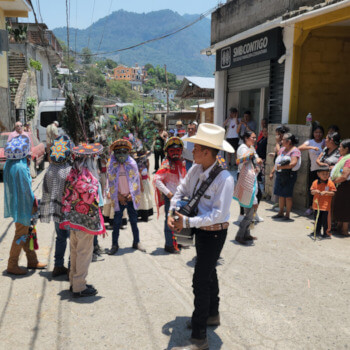 Thumbnail image of a festive street in rural Mexico, showing people in traditional costumes and masks dancing, with a young man playing guitar in the center.